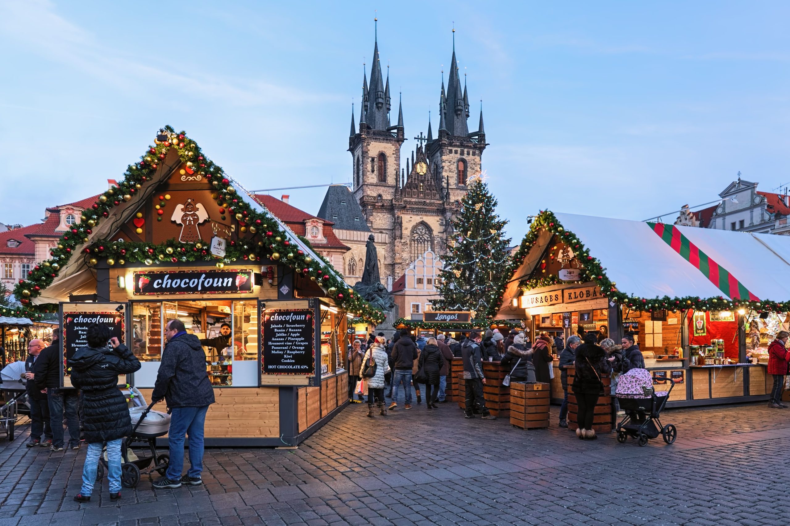 Festive Prague Christmas Market in Old Town Square with decorated wooden stalls, a large Christmas tree, and the Church of Our Lady before Týn in the background at dusk.